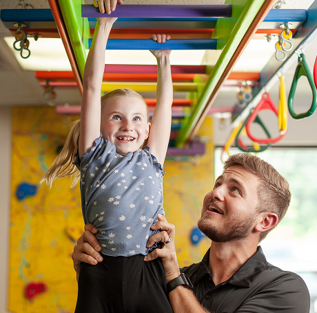 Pediatric therapist supporting a young girl as she reaches across colorful monkey bars